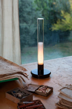 Sylvestrina portable lamp on a table by a window, beside papers and a box of pastels