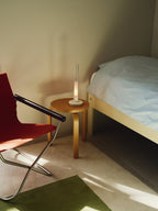Sylvestrina portable lamp on a bedside stool beside a red folding chair